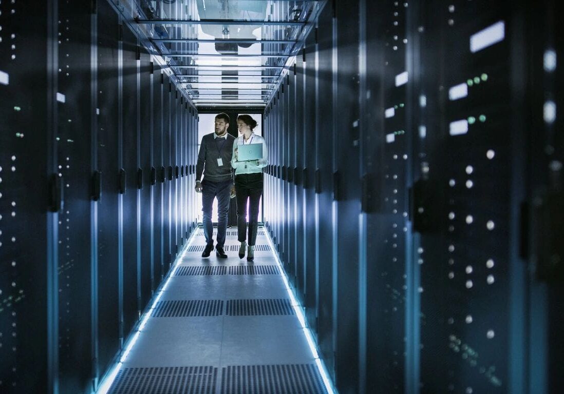 Two people are walking through a hallway of servers.